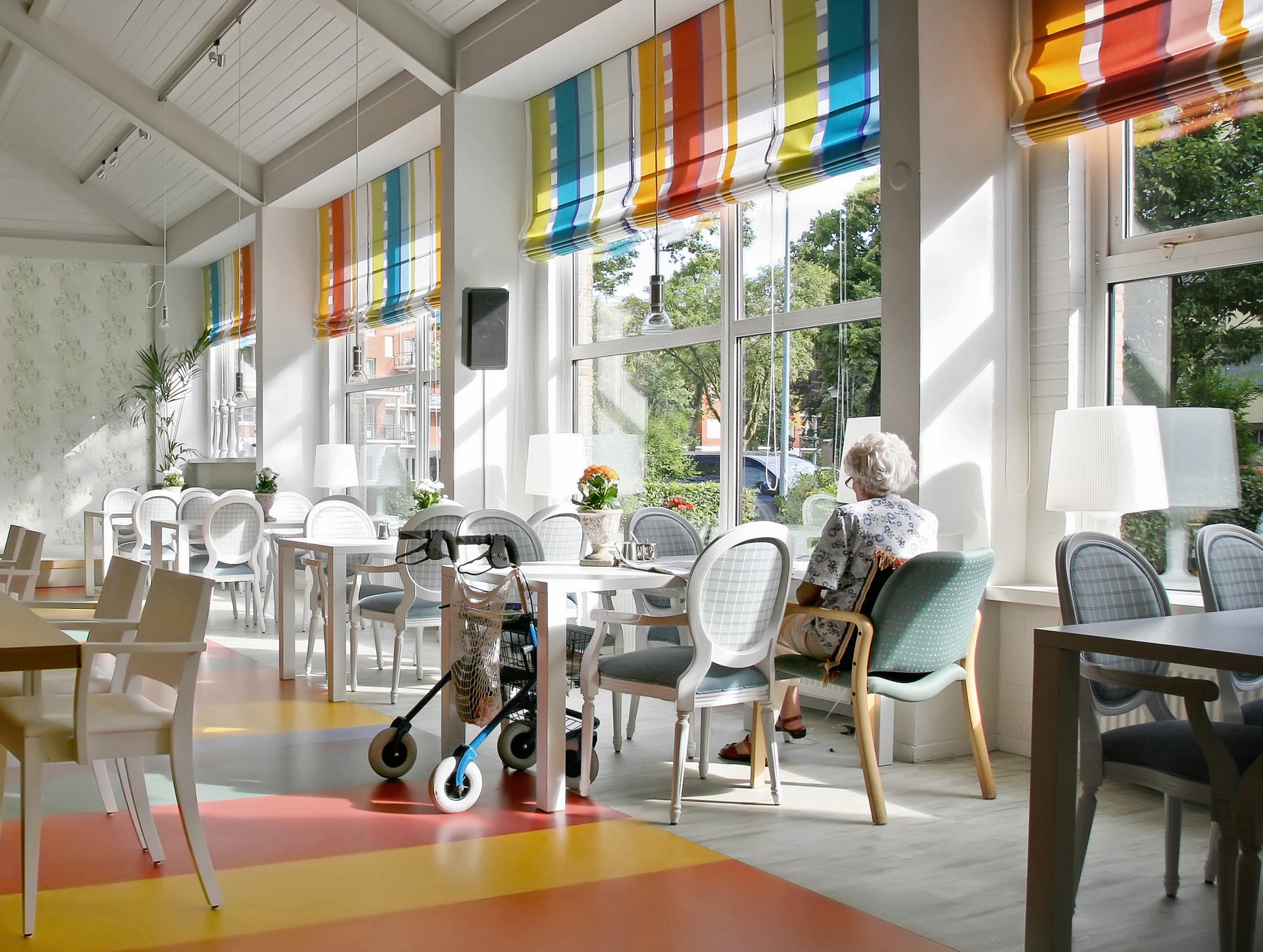 Bright, modern care home dining lounge with floor-to-ceiling windows, colourful blinds, and a resident seated by the window with a rollator nearby