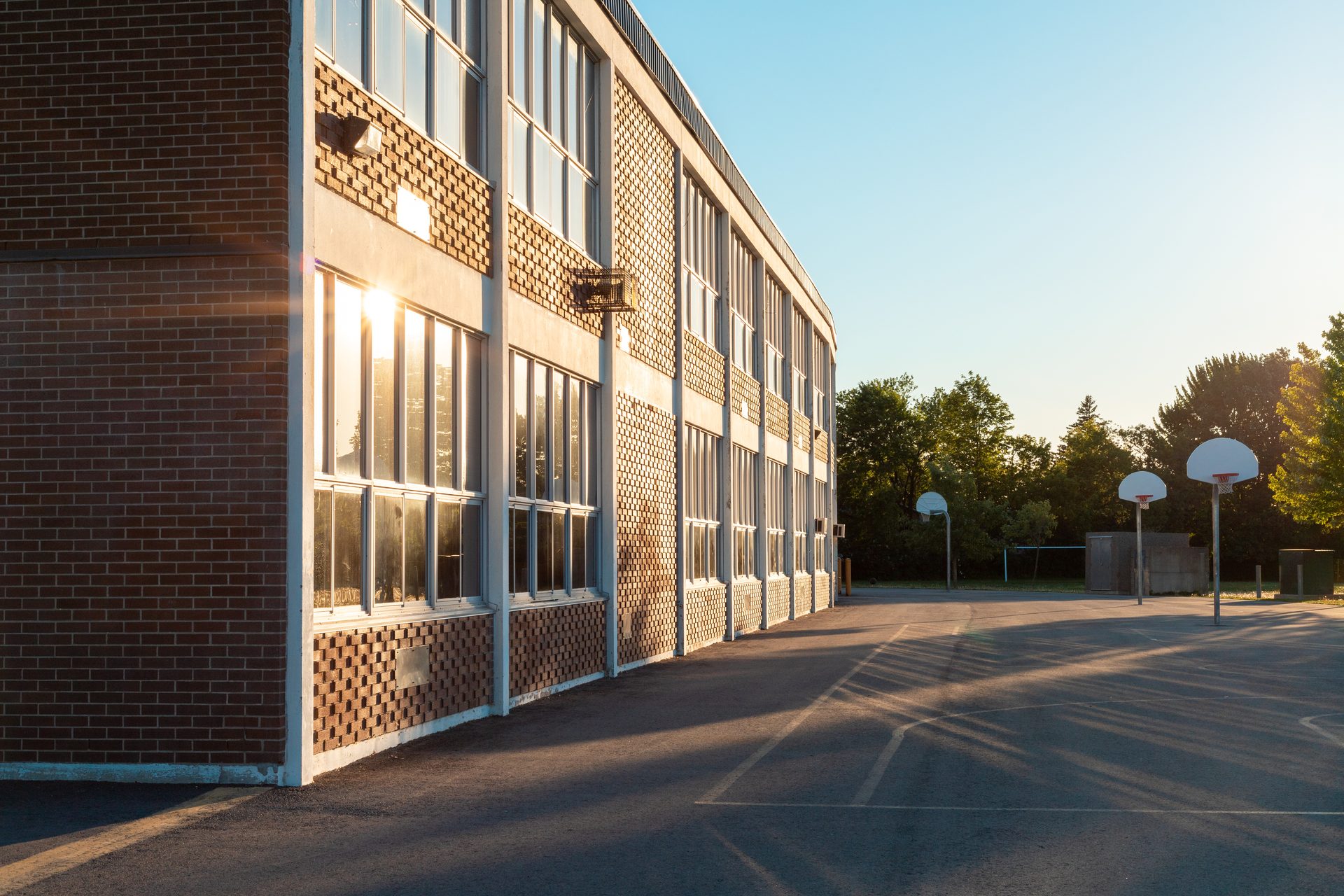 School building exterior at golden hour, with empty playground and basketball hoops, conveying the out-of-hours stillness of the holiday inspection window