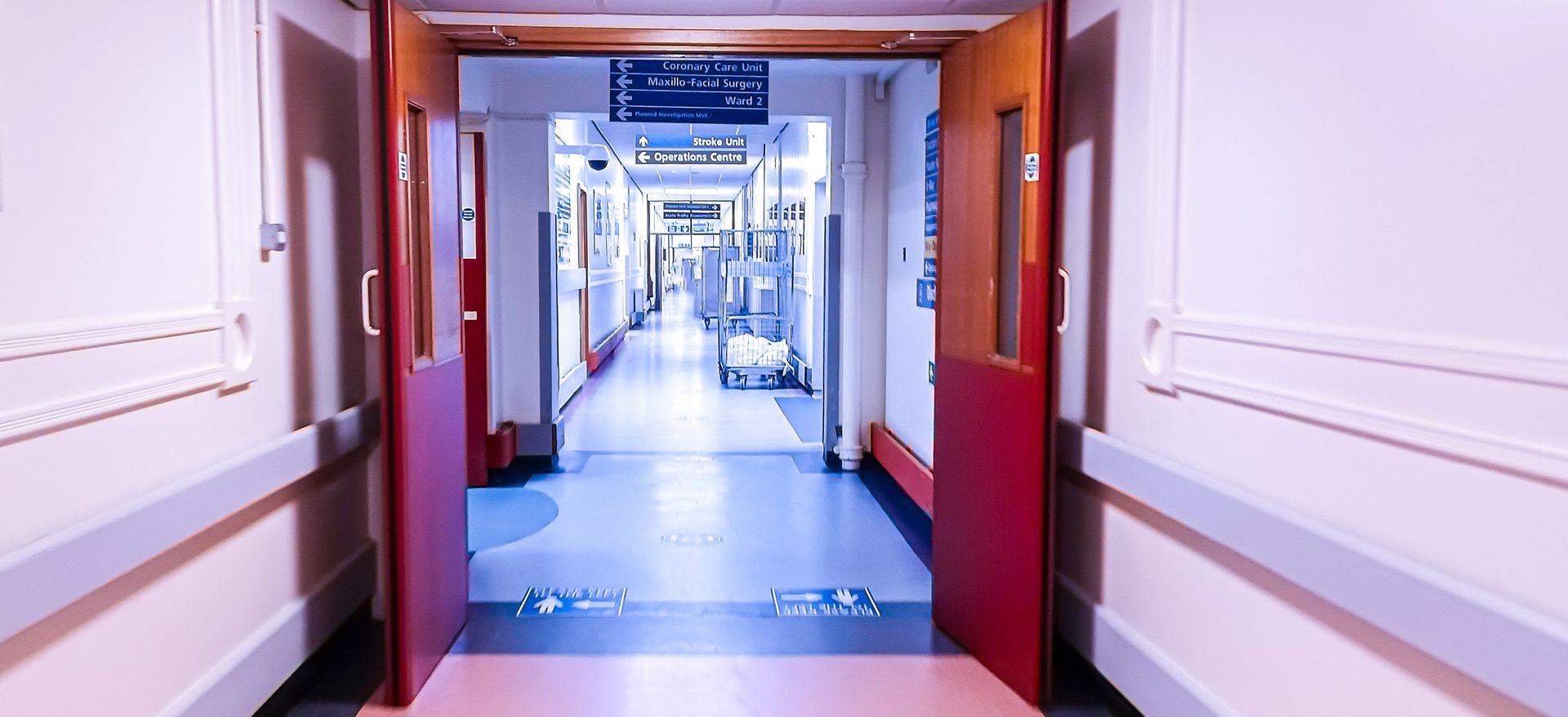 View through a pair of open red fire doors into an active NHS hospital corridor with ward signage and a linen cart