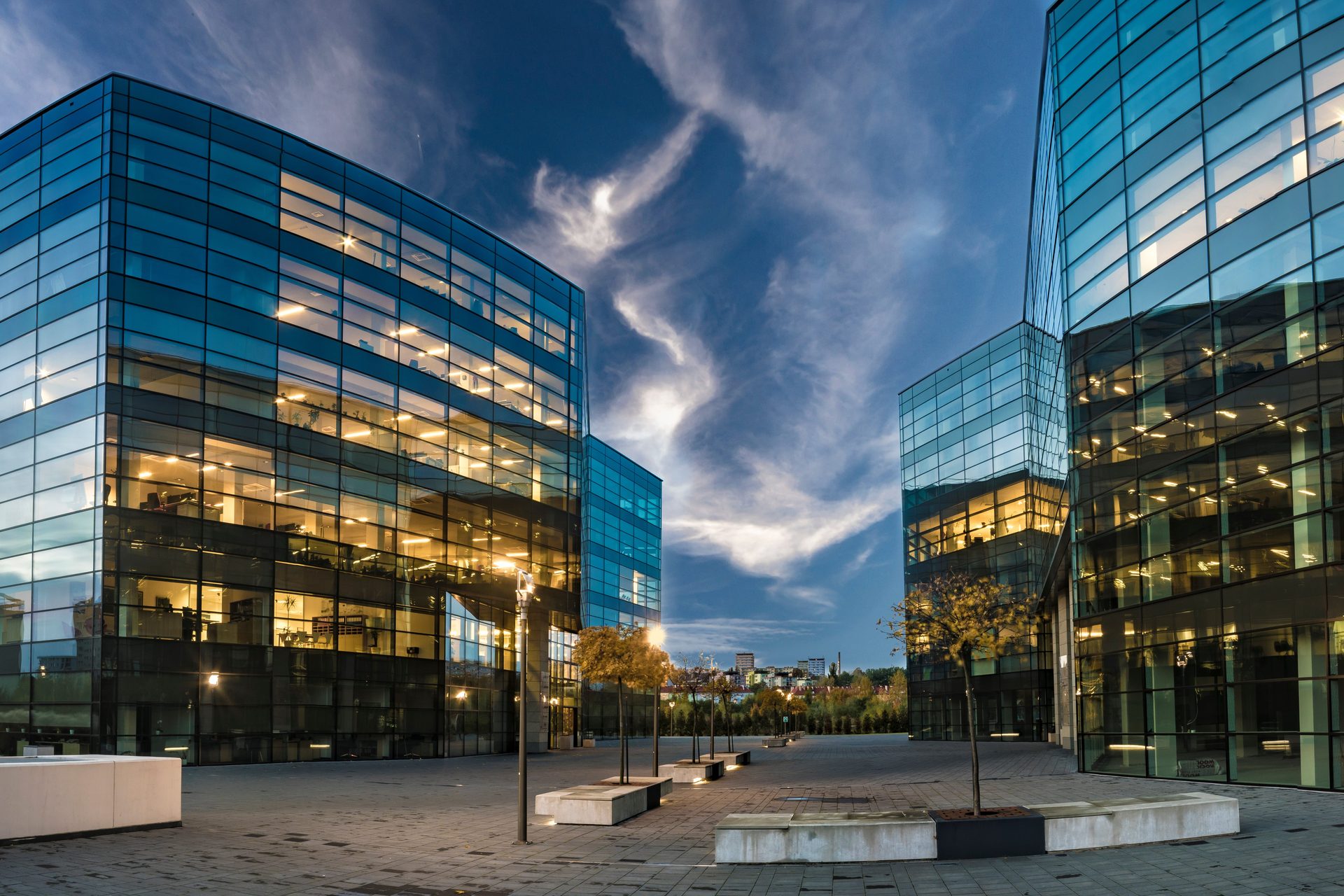 Two modern Class A commercial office buildings at dusk, glass facades illuminated from within, plaza and landscaping between them