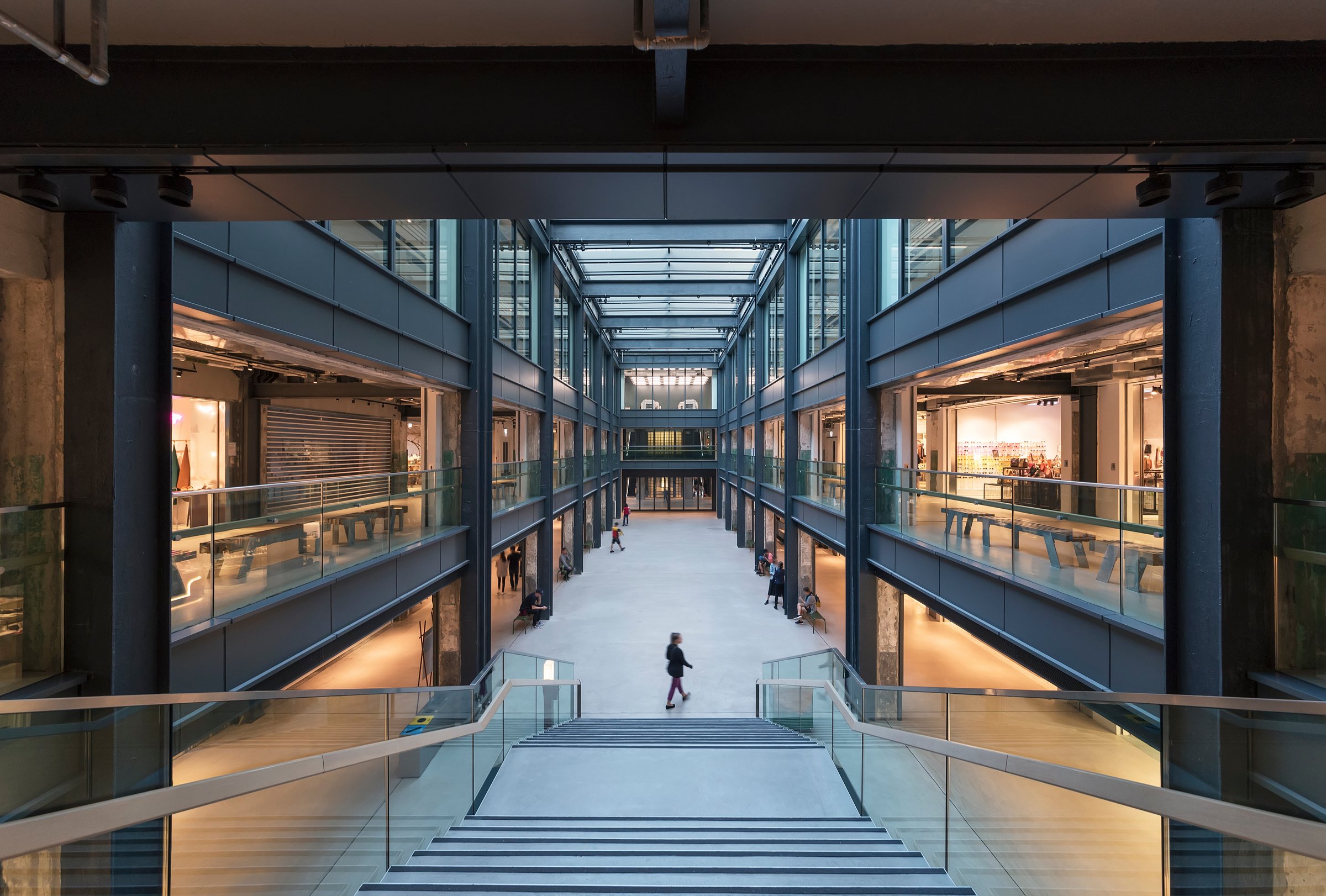 Modern industrial-style shopping centre atrium with multi-level retail units, steel-framed walkways, glass balustrades, and shoppers walking through the central concourse